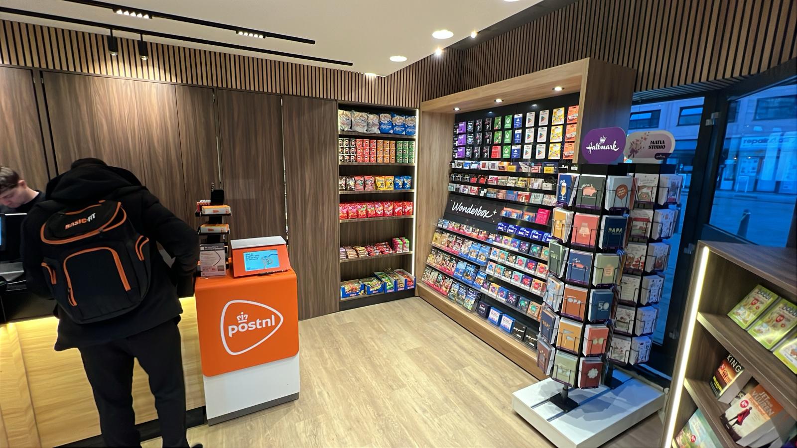Gerealiseerde kiosk Rotterdam — PostNL-balie met houten lattenwand, verlichte snackschappen en Hallmark wenskaartendisplay