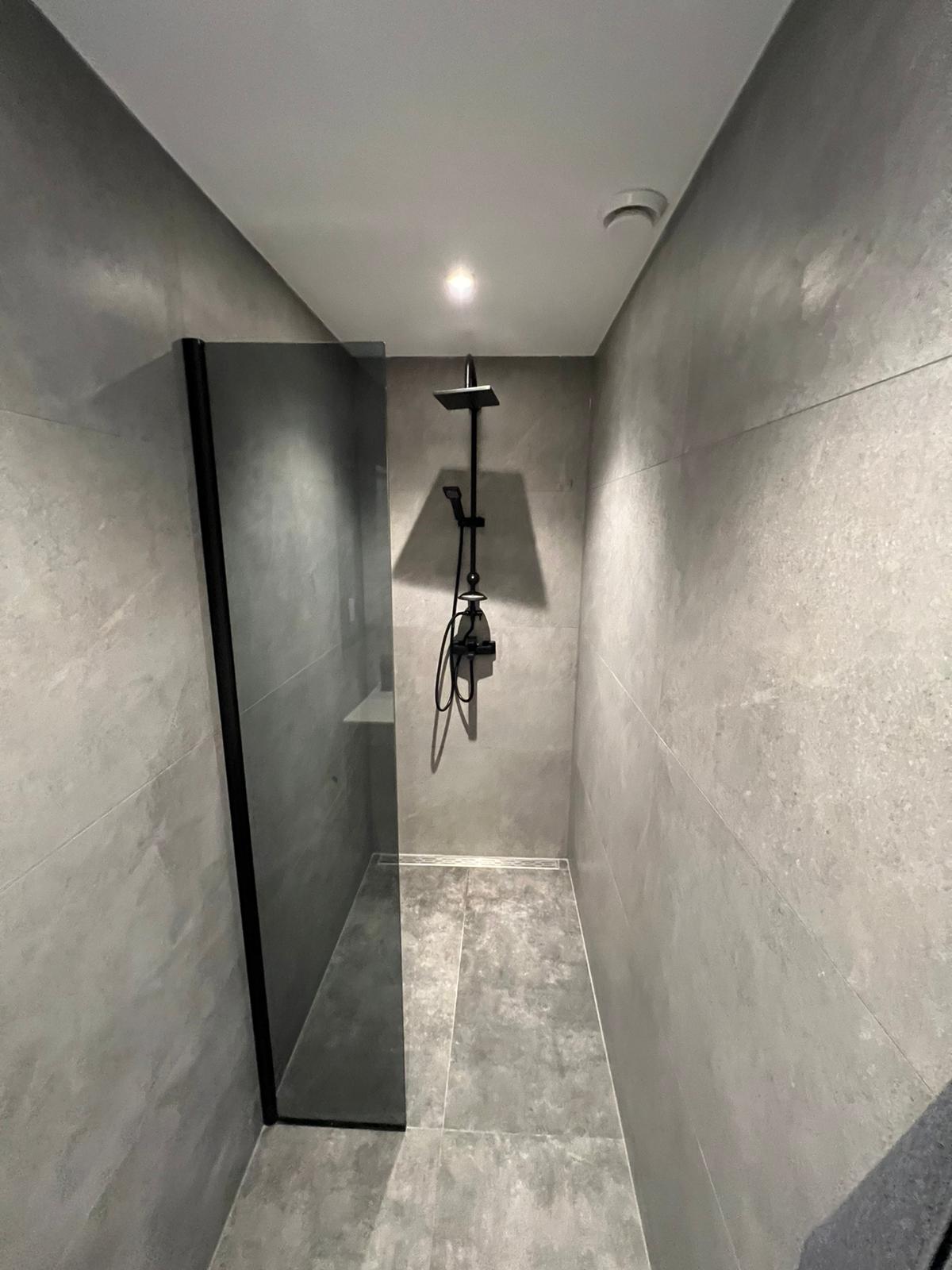 Inloop­douche met groot­formaat beton-look tegels, mat-zwarte regen­douche en zwart glazen wand
