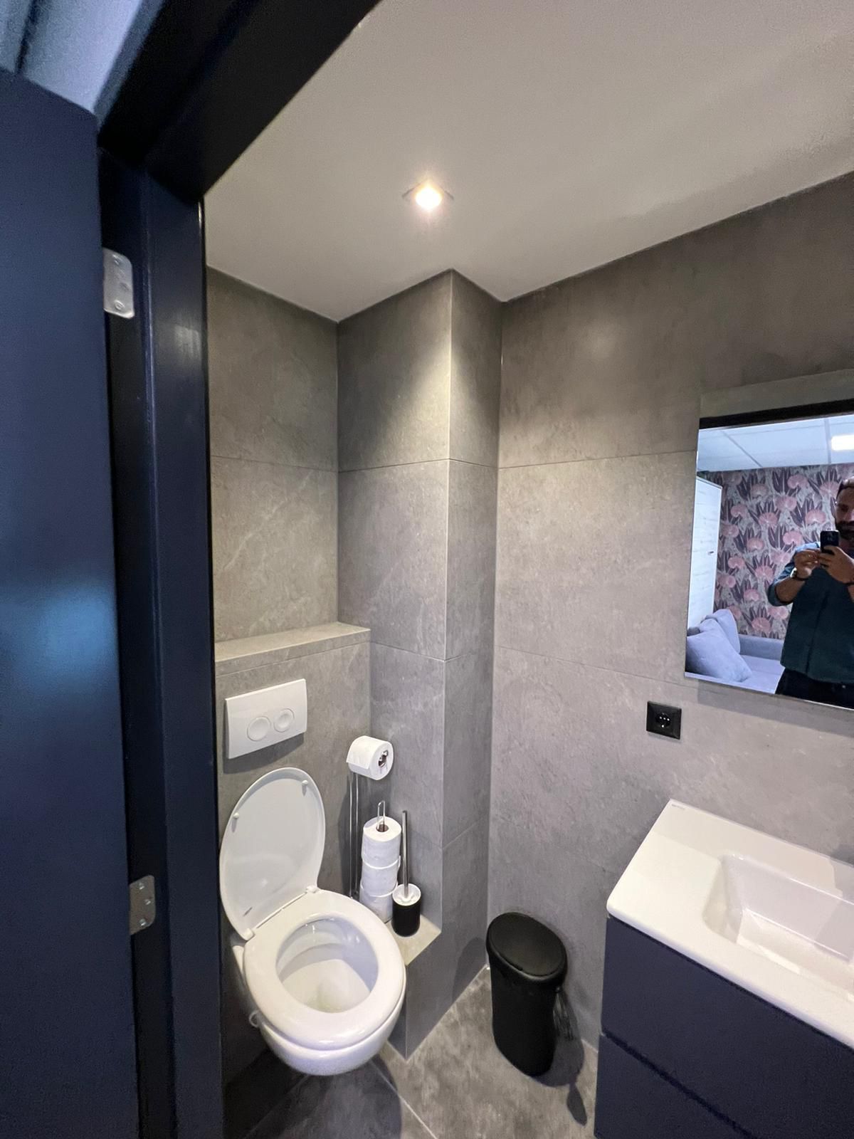 Toilet­ruimte met hangtoilet, witte druk­plaat, kleine wastafel en grijs tegelwerk